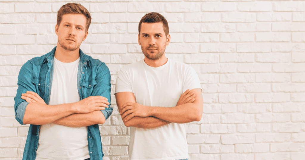 Two men stand side by side against a white brick wall with arms crossed and guarded expressions, illustrating how being measured against someone else quietly humiliates a narcissist who needs to feel superior.