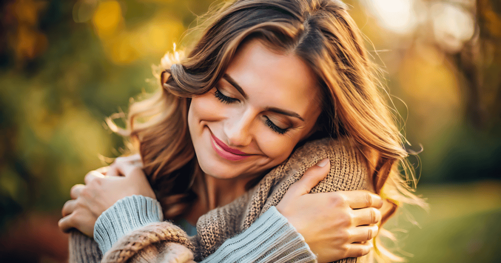 A smiling woman wraps her arms around herself in a gentle, warm embrace outdoors, illustrating the comforting self-compassion that restores inner peace.