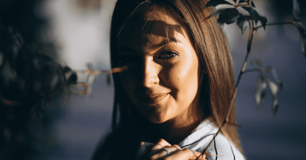 A woman smiles softly from behind leaves with warm sunlight grazing her face, reflecting the serene clarity that comes from finally releasing unwarranted self-blame.