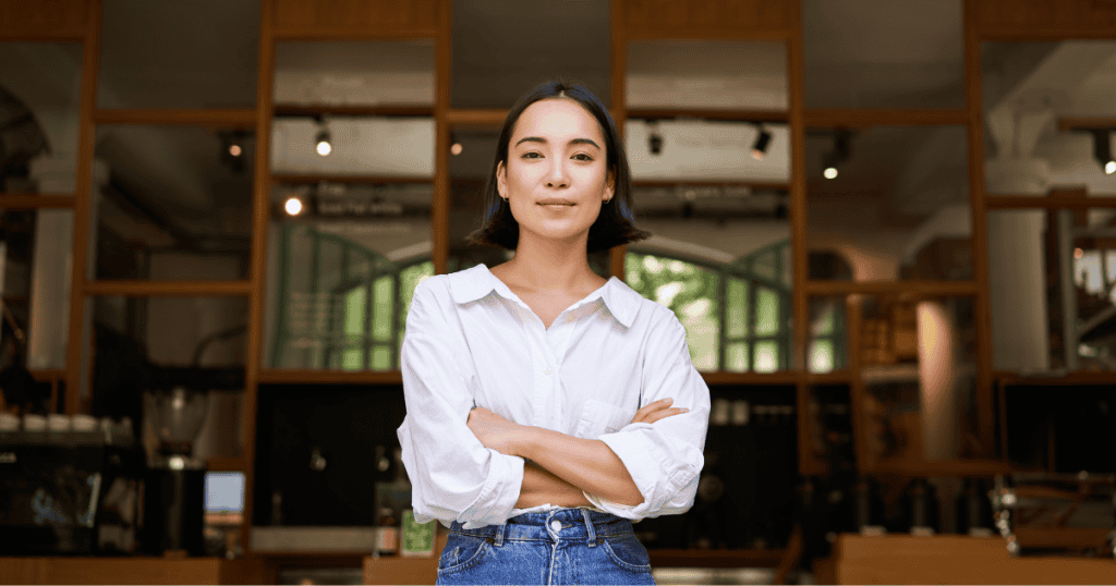 A young woman in a crisp white shirt stands with crossed arms and a steady, direct gaze inside a modern cafe, portraying the quiet confidence of holding firm personal boundaries.