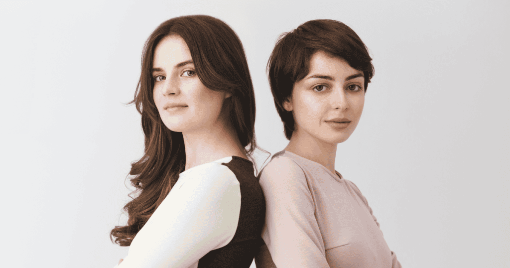 Two young women stand back-to-back with guarded, reserved expressions against a stark white backdrop, depicting the subtle division and competition manufactured between family members.
