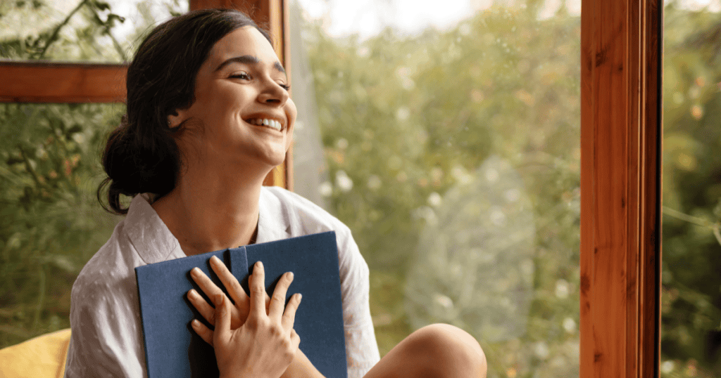 A woman laughs joyfully by a sunlit window while holding a journal close to her chest, embodying the relief and clarity of finally knowing the truth.