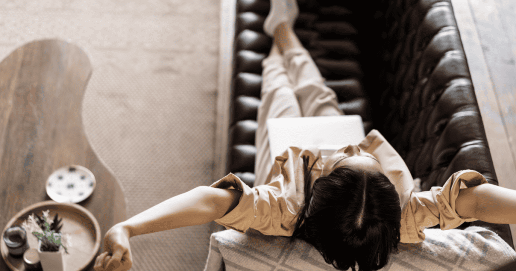 An overhead shot shows a woman lying back on a couch with arms outstretched, eyes closed in total relaxation, embodying the version of herself who finally allows rest.