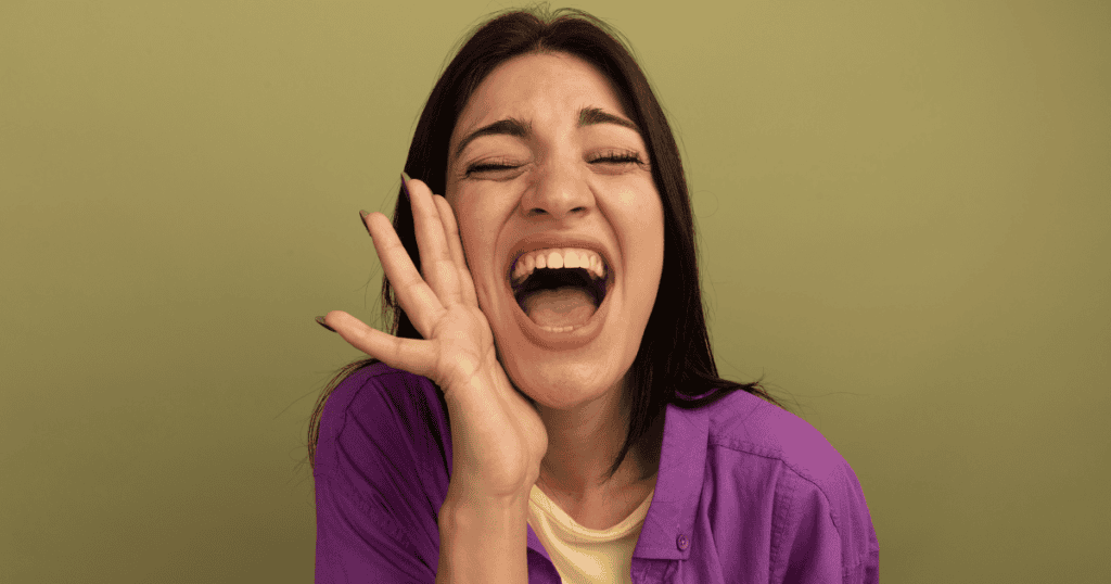 A woman in a purple shirt throws her head back and laughs joyfully with a hand raised to her cheek, depicting the exuberant freedom of seeing through the game and breaking out for good.