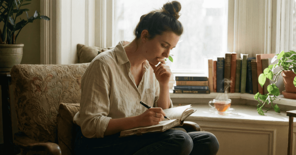 A woman sits curled comfortably in an armchair journaling beside a sunlit bay window, embodying the quiet, grounding peace of cultivating your own inner sanctuary.