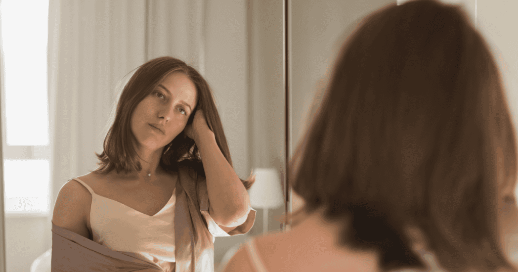 A woman in a silk robe looks thoughtfully at her reflection while tucking hair behind her ear, capturing a gentle, grounded moment of self-reflection and re-centering.
