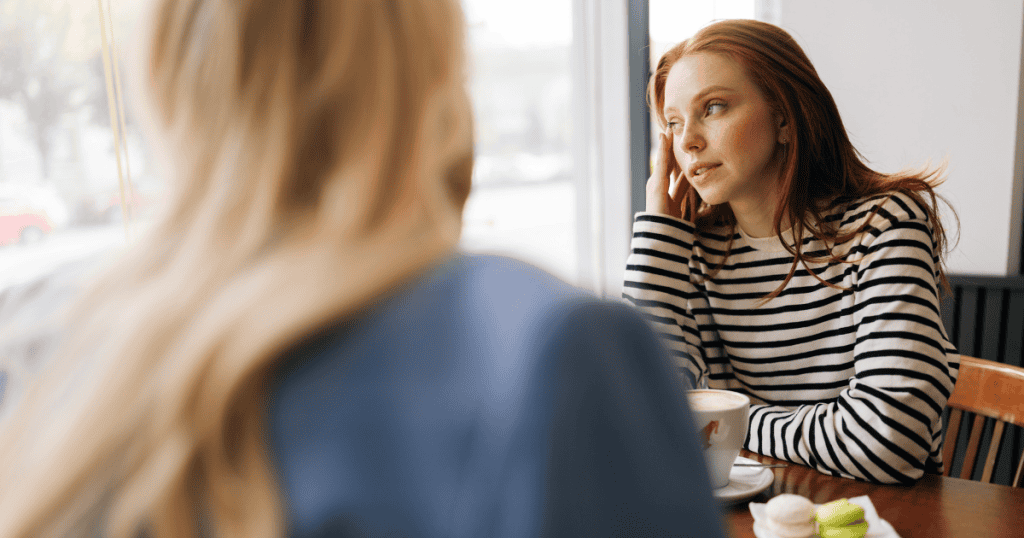 A red-haired woman rests her temple against her hand while listening intently across a cafe table, portraying the careful discernment needed when navigating guarded conversations.