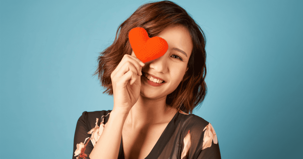 A smiling woman playfully holds a small knitted red heart over one eye against a bright blue studio backdrop, symbolizing the blinding illusions that disguise emotional need as love.