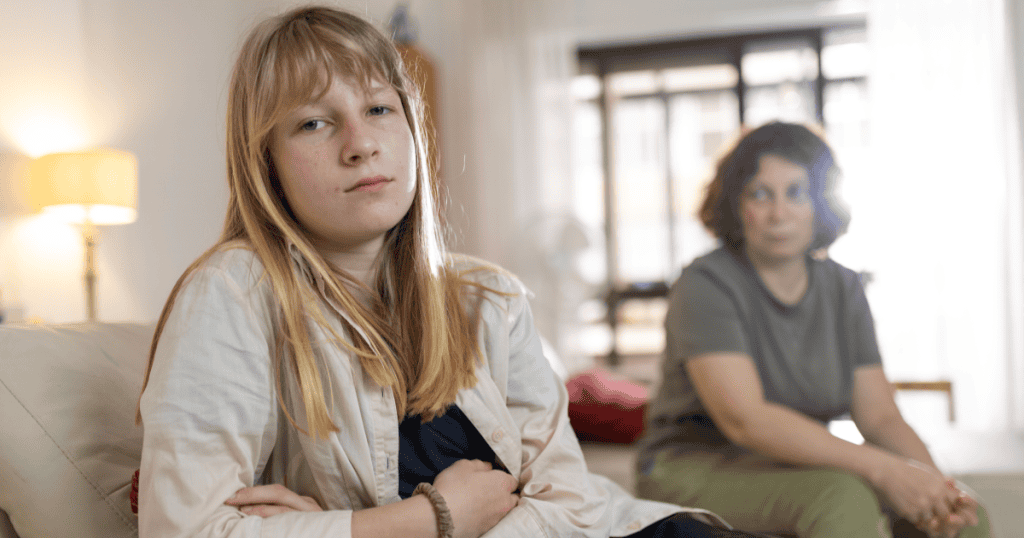 A young woman sits with a guarded, unreadable expression while an older woman watches from the background, capturing the quiet toll of intimacy that's consistently withheld.