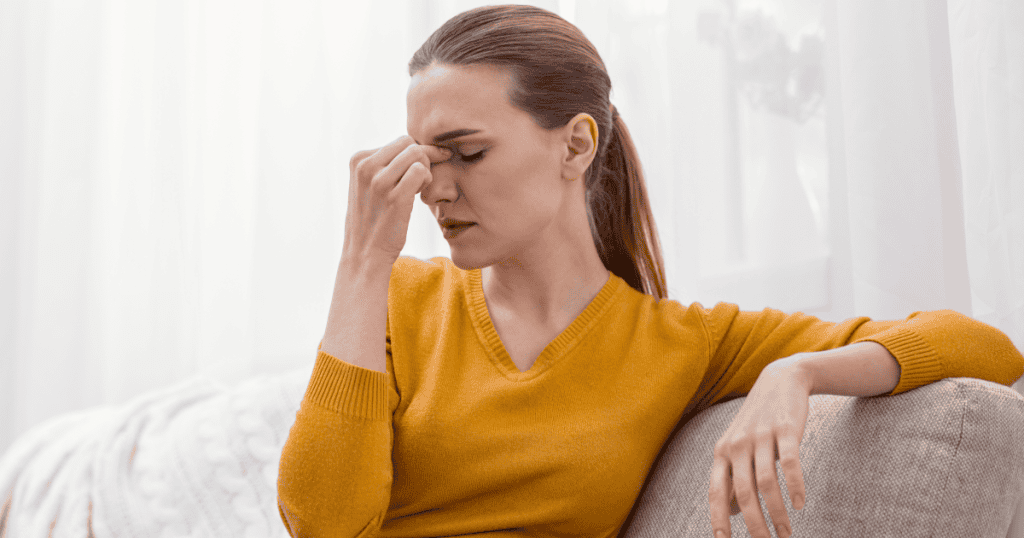 A woman clutching her head in pain on the couch, showing how a narcissist's psychological abuse doesn't stay in the mind, but manifests as real, physical stress and exhaustion in the body.