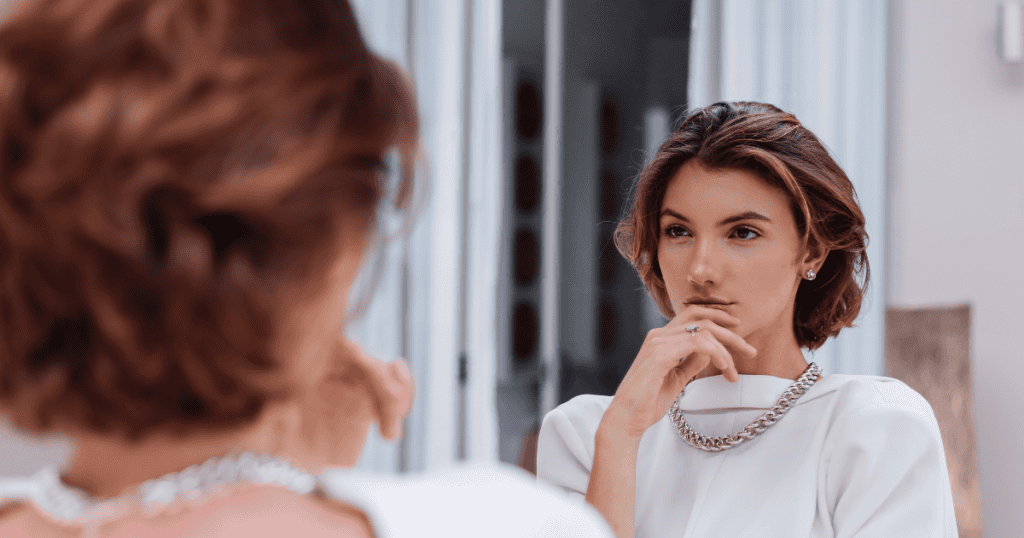 A poised woman in an elegant white blouse rests her fingers near her chin while looking steadily at another woman across from her, reflecting the composed, observant presence of holding firm personal boundaries.