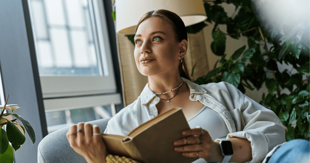 A woman reading a book by the window with plants around her.