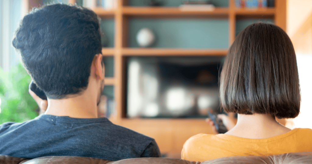 A couple sits side by side facing away from the camera watching TV together, their turned backs symbolizing the emotional distance kept just beneath the surface.