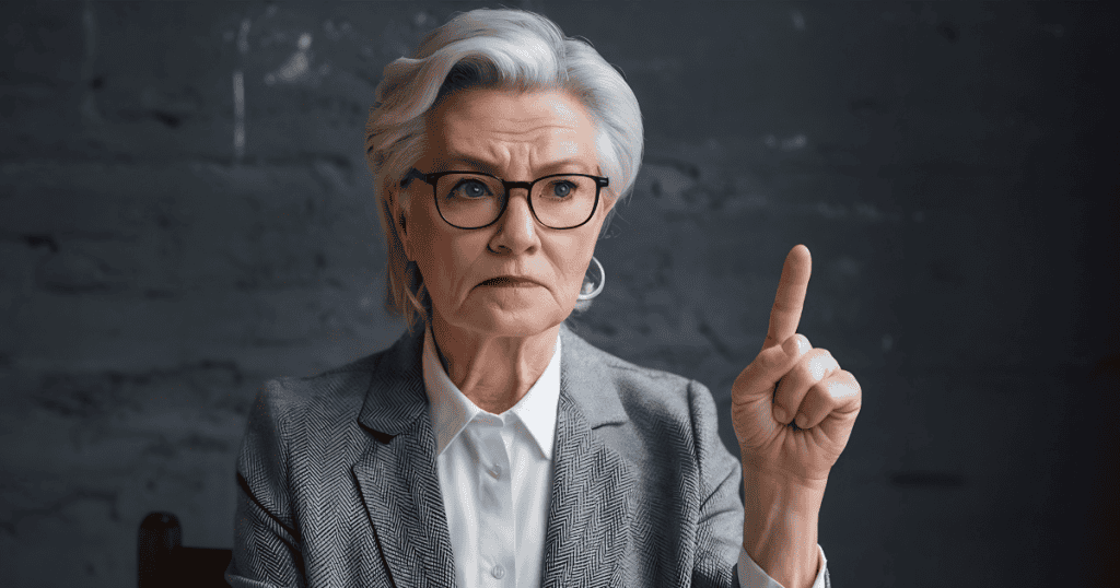 A severe woman in a grey tailored blazer raises a stern warning finger with a grim frown, showcasing the unyielding authority used to enforce compliance and suppress individual voices.