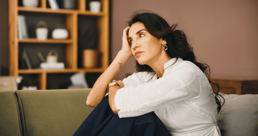 A woman leans her head against her hand on a sofa with a distant, wounded gaze, embodying the quiet emotional erosion caused by constant nonverbal hostility.