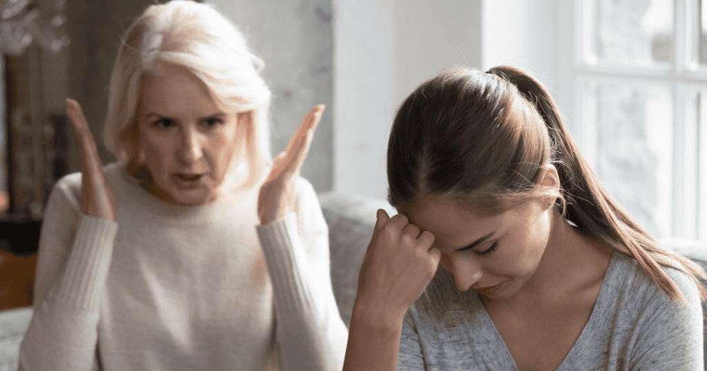 An older woman gestures angrily with raised hands while a younger woman bows her head in distress, depicting the exhausting emotional weight of enduring chronic, volatile blame.