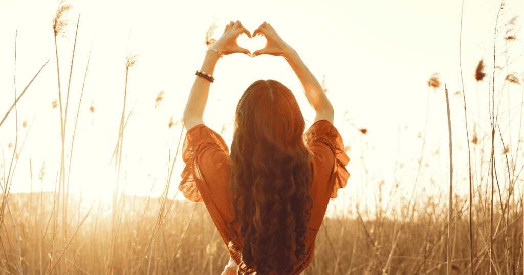 A woman viewed from behind frames the glowing sunset with her hands in the shape of a heart amidst tall field grasses, symbolizing the liberating warmth of reclaiming love for oneself.