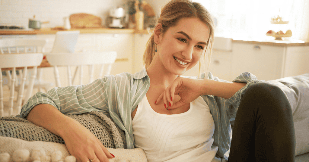 A relaxed woman smiles with easy radiance while lounging back comfortably on a sunny sofa, embodying the restorative peace of knowing your worth was never the issue.