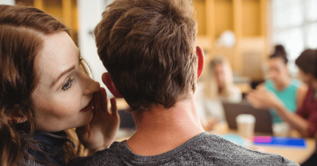 A woman leans in to whisper secretly into a man’s ear while others work nearby, illustrating the hidden things a narcissist says about you when they believe it will never reach you.