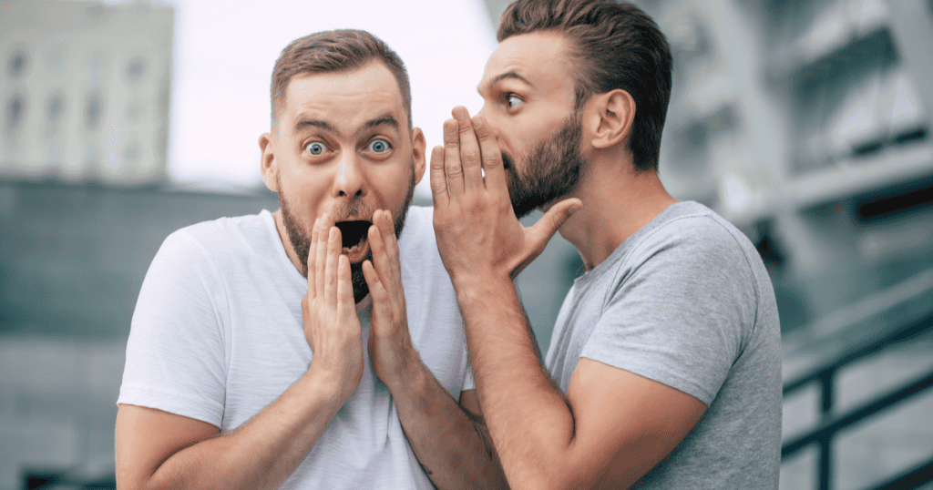 A man whispers into his friend’s ear as the friend covers his mouth in shock, capturing the kind of damaging talk a narcissist spreads about you to others behind your back.