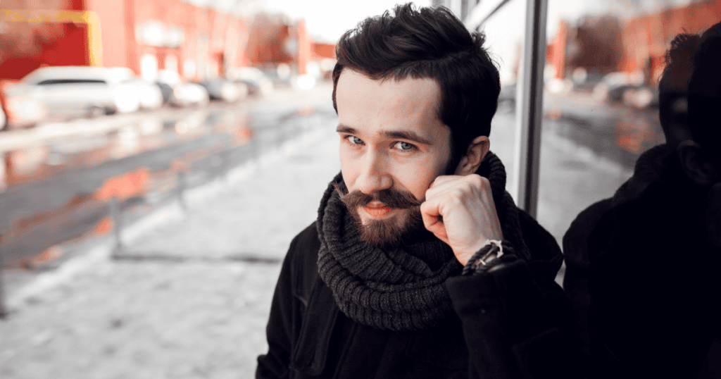A bearded man in a scarf leans on a window with a sly, knowing smirk, hinting at the sneaky tests a narcissist uses to check whether they still have control over you.