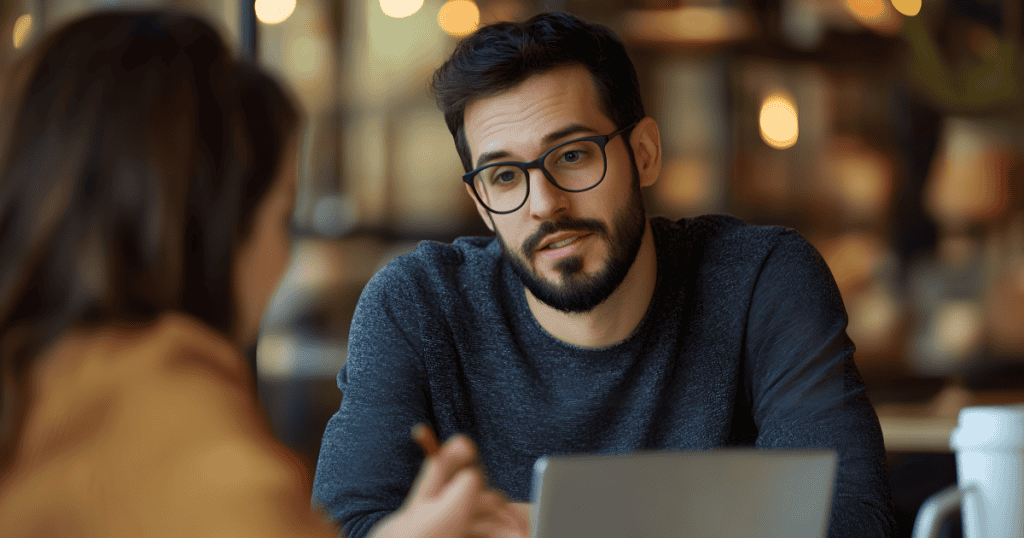 A bearded man in glasses speaks calmly across a café table with a subtly dismissive look, illustrating how a narcissist’s quiet, toxic phrases can wear down your confidence without you noticing.