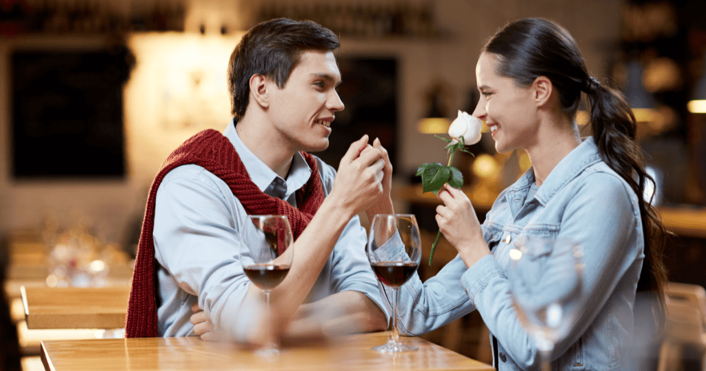 A smiling woman holds a white rose while a charming man clasps her hand over glasses of red wine, illustrating how a narcissist's romantic early attention can feel so genuine that you don't realize who you're dating.