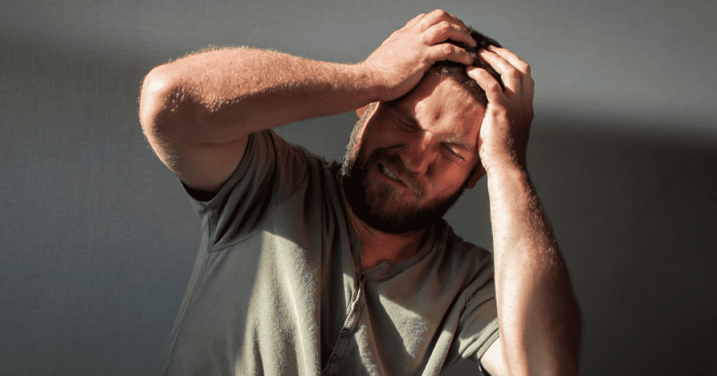 A bearded man clutches his head with both hands and grimaces in frustration, illustrating how easily a narcissist’s fragile ego can be wounded without you even trying.