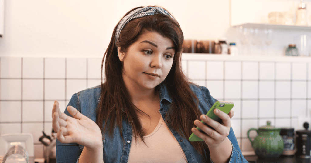 A woman looks at her phone with a baffled expression and an open-handed shrug, capturing the frustration of being told to “calm down” by a narcissist who dismisses your feelings.