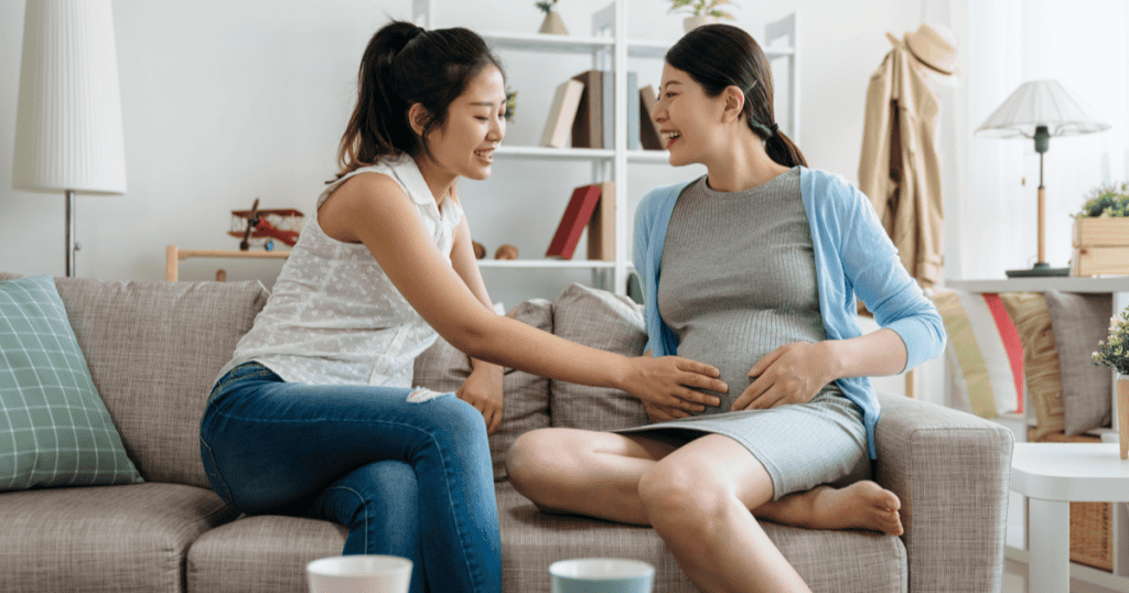 A smiling woman rests her hand on her pregnant friend’s belly as they laugh together, illustrating how a narcissist’s warm gestures can later be held over you as debts you owe them.