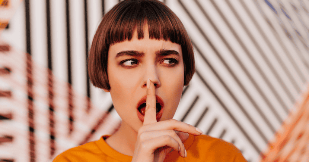 A woman holds a finger to her lips with a sideways glance, showing the power of staying quiet instead of explaining or justifying yourself to a narcissist.