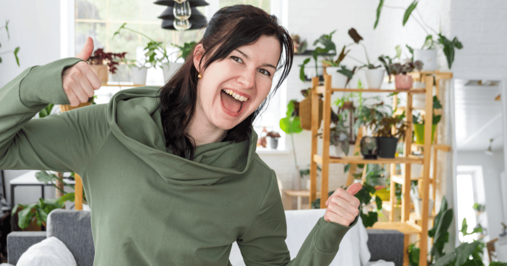 A woman gives two enthusiastic thumbs up with a huge smile in her plant-filled home, celebrating how building a happy life of your own is the only payback you need after a narcissist.