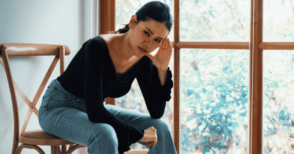 A woman sits by a window with her hand to her head and a weary, drained expression, capturing the emotional toll of living alongside a narcissist’s carefully maintained image.