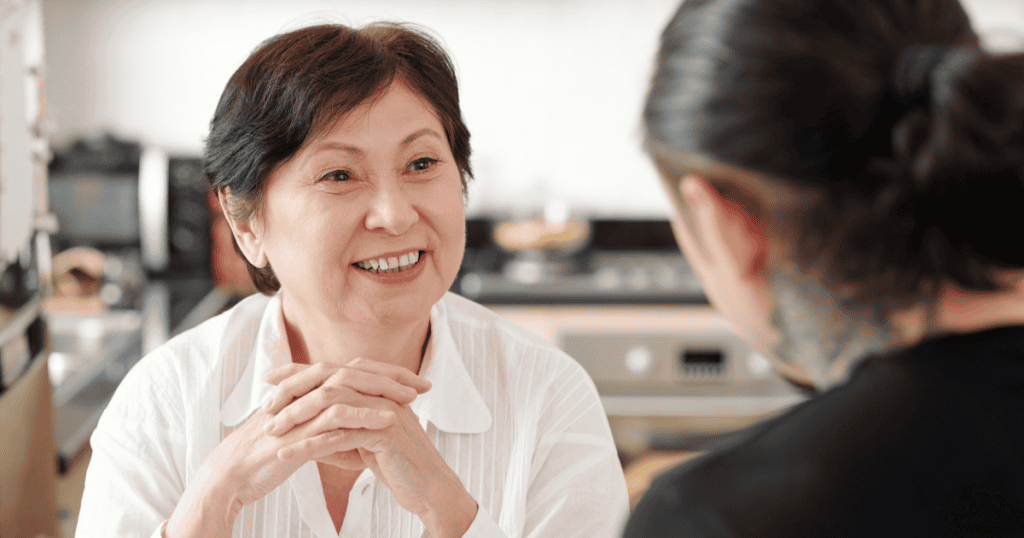 An older woman smiles warmly with her hands clasped as she talks with a younger woman, illustrating how a narcissist hides small tests of exclusion behind friendly conversation to see how you react.