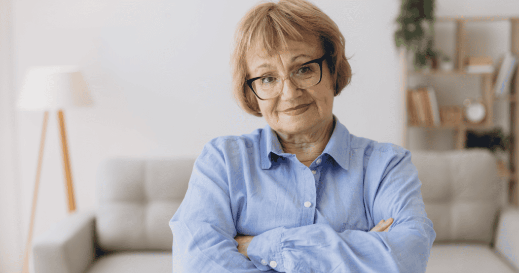 An older woman in glasses stands with arms crossed and a smug half-smile, reflecting how a narcissist takes credit for your success by claiming you wouldn’t be where you are without them.