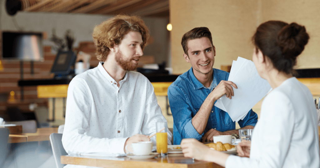 Two men lean in eagerly across a café table, one smiling as he holds out papers to a woman, illustrating how a narcissist starts working hard to win back your attention once you stop chasing them.