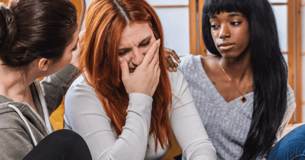 A red-haired woman covers her mouth as she cries while two people comfort her, illustrating how a narcissist's tearful return can trigger your empathy and pull you back into caring for them.