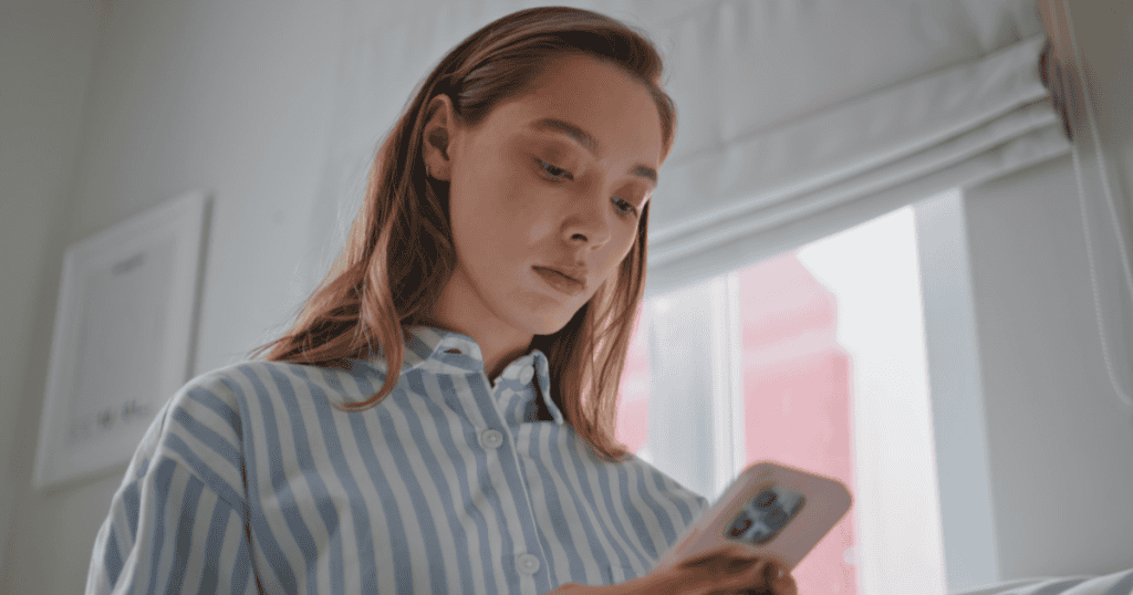 A young woman looks down at her phone with a wary, unimpressed expression, capturing the moment of seeing a narcissist's message and recognizing their return was never about genuinely missing her.
