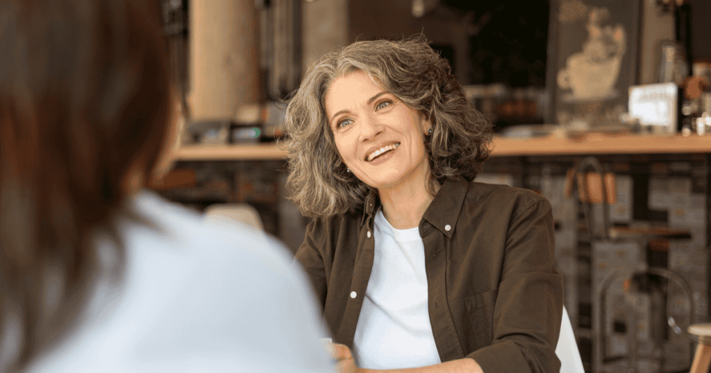 A gray-haired woman smiles attentively at someone across a café table, illustrating how a narcissist’s warm, engaged manner lets them read you with unsettling speed.