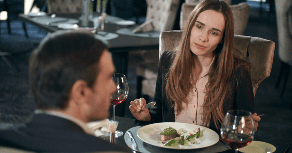 A woman listens with a polite, uncertain smile as a man talks across the table at an elegant dinner date, showing how the pattern of choosing narcissists can quietly repeat itself without you noticing.