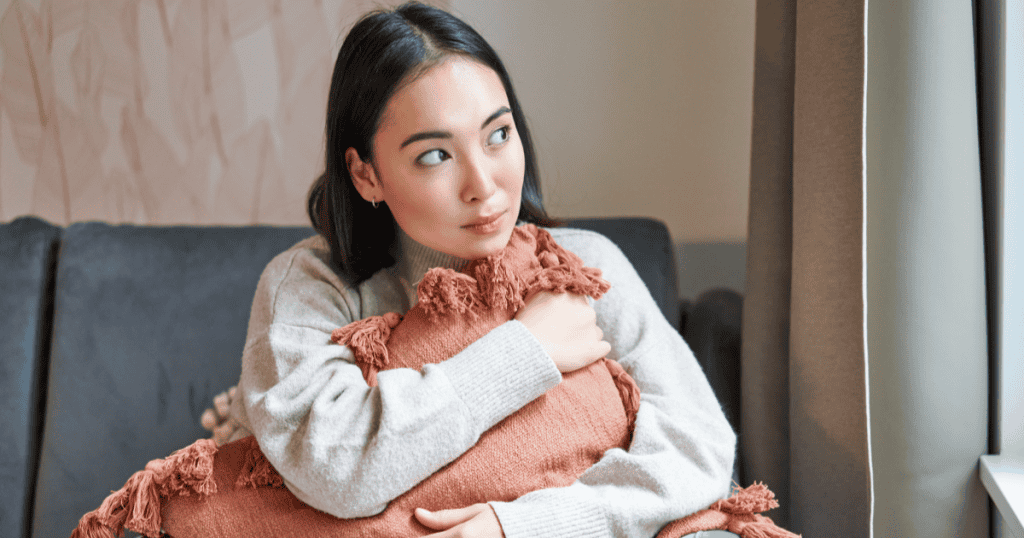 A young woman hugs a cushion on the sofa and gazes thoughtfully out the window, reflecting on how her genuine presence disrupted something in a narcissist that they could never quite explain.