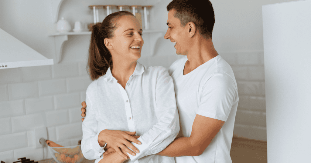 A woman laughs in the warm embrace of her partner in a bright kitchen, showing the ease of being with someone who accepts you without asking you to keep apologizing for who you are.
