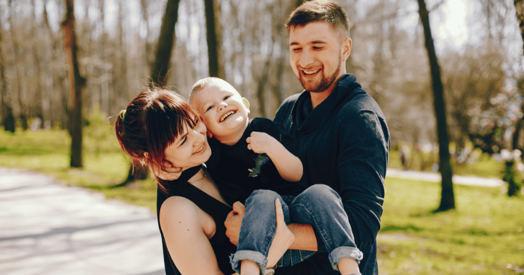 A smiling couple holds their laughing toddler on a sunny park path, showing the joyful life you can build once it no longer revolves around a narcissist.