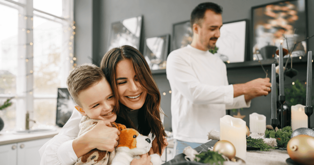 A smiling woman hugs her young son while a man decorates the table in a warm, festively lit home, showing the full and joyful life you can build even while remaining an unfinished chapter in a narcissist's story.