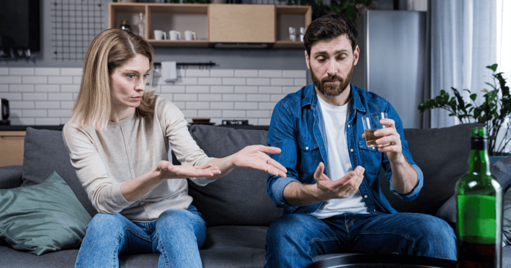 A frustrated woman gestures toward a man holding a glass of whiskey on the couch beside a liquor bottle, illustrating how you may keep being drawn to partners who are using, a pattern that can lead you into relationships with narcissists without realizing it.