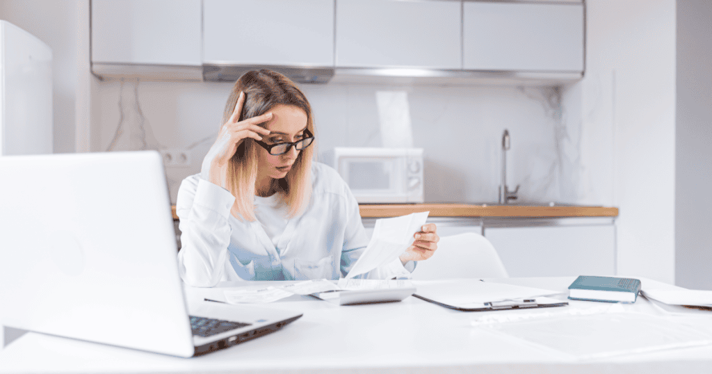 A woman frowns over bills and a calculator at her kitchen table, reflecting how a narcissistic parent deliberately left their child unprepared for adult life so they would stay dependent.