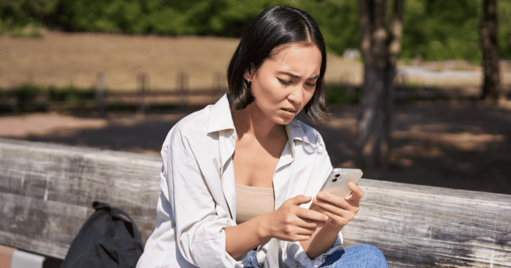 A woman sitting on a park bench frowns uncomfortably at her phone, capturing the unease of being tagged in an old photo by a narcissist testing whether they can still get a reaction from you.