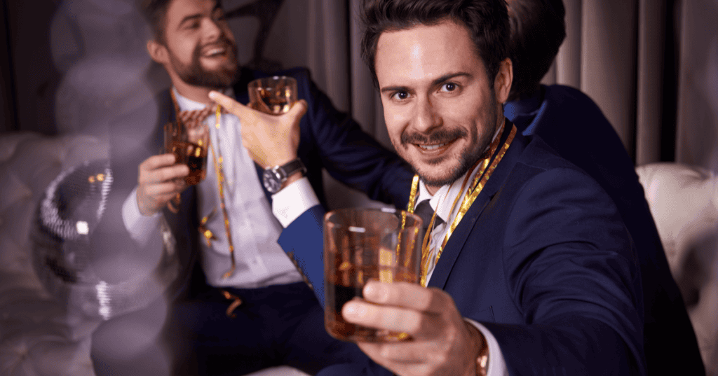 A polished man in a suit raises his whiskey glass toward the camera with a charming smirk at an upscale party, illustrating how a narcissist's more refined, careful persona around money and status is still just another mask.