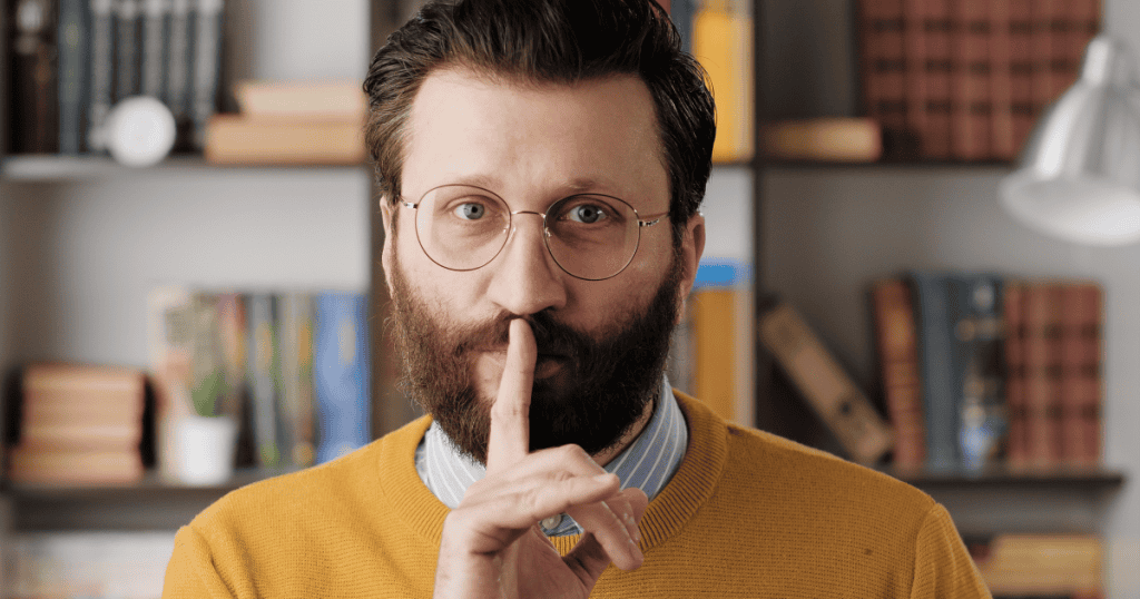 A bearded man presses a finger to his lips in a hushing gesture, illustrating how a narcissistic partner keeps you away from their inner circle so their secrets never come to light.