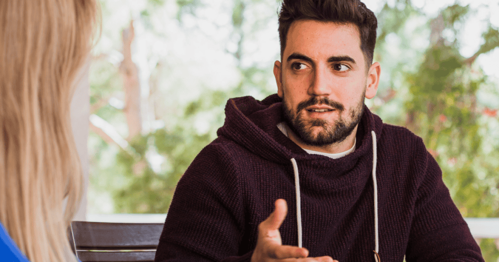 A man in a hoodie gestures casually while watching a woman’s reaction closely, illustrating how a narcissist drops a meaningful name into conversation to see what emotions it stirs in you.
